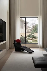 A minimalist living room with a textured cloth recliner chair, a flat-screen television, and a large window with white curtai