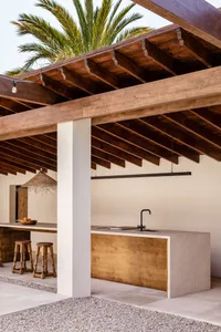 A modern exterior kitchen and dining space featuring wooden beams, concrete counter, and palm trees.