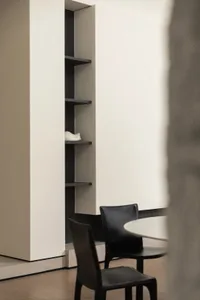 Two black chairs with a round table in front of a minimalist shelving unit with wood shelves and a white wall background.