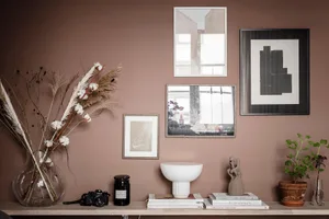 A minimalist home library with a ceramic bowl, books, and plants on a wooden surface against a mauve-colored wall with framed