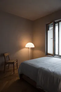 A minimalist bedroom with a wooden floor and fabric bedding, an open window with blinds, and a floor lamp illuminating the sp