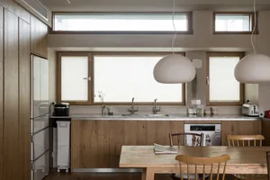 A minimalist residential kitchen featuring wood and stainless steel elements, with ample natural light through large windows.
