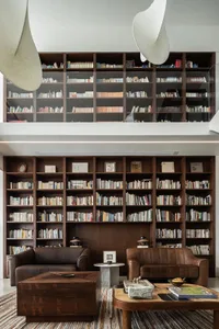 A modern reading room featuring wooden shelves filled with books, two brown leather sofas, and a wooden coffee table with var