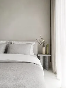 A minimalist bedroom featuring a bed with gray and white textiles, a side table with plants and vases, and white curtains.