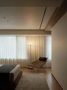 Minimalist bedroom featuring a wooden bed, glass windows, and fabric curtains, illuminated by a modern floor lamp.