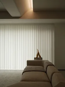 A minimalist living room with a taupe-colored sofa, fabric blinds, and a small gold-colored sculpture on a shelf.