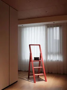 Red metal step ladder with a black purse on it positioned against a white curtain in a modern home interior.
