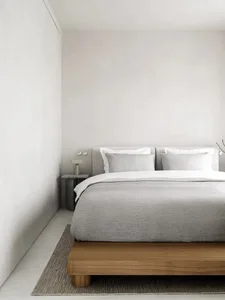 Minimalist bedroom featuring a wooden bed platform, gray and white bedding, and a textured side table with a lamp.