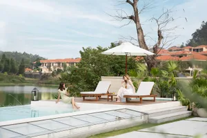 Two women sitting on wooden lounge chairs under a large white umbrella, enjoying drinks by a pool with a view of surrounding