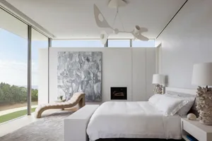 A minimalist bedroom with modern white furniture, large windows, and abstract artwork.