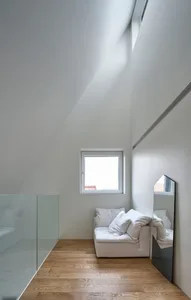 A minimalist white canvas sofa situated under a window in a room with a sloped ceiling and glass barrier.