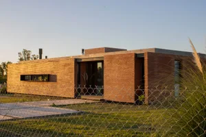 Modern house with brick exterior and large windows, surrounded by greenery and a gravel road.