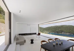 A modern minimalist living room with a white concrete floor and walls, featuring a large window with a view of lush green hil