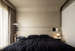 A minimalist bedroom featuring black textiles on the bed, a textured beige headboard, and neutral-toned curtains.