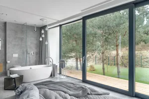 Modern bathroom with concrete walls, glass shower enclosure, and a large window overlooking a forested area.