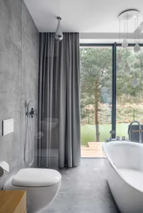 A modern bathroom featuring a concrete shower, gray curtains, and a bathtub overlooking a forested area.
