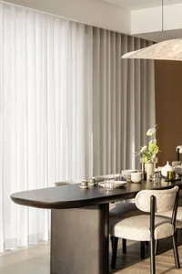 A modern minimalist dining room featuring a sleek, dark wooden table and beige upholstered chairs, surrounded by sheer curtai