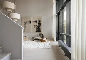 A modern, neutral-toned living room with minimalist decor, light-colored wood floors, and large windows.