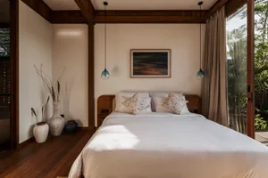 A minimalist bedroom with a wooden bed and white bedding, featuring a painting and blue pendant lights.