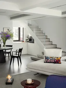 A minimalist living room with white decor, featuring a comfortable sofa, a round table, black chairs, and a glass staircase.
