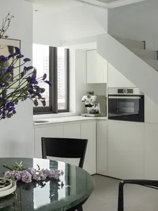 A modern kitchen featuring white cabinets, a black marble table with purple flowers, and a window with a cityscape view.