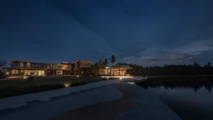 A luxurious waterfront villa featuring glass walls, wooden accents, and stone flooring, illuminated by warm exterior lighting