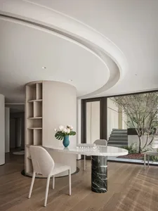 A modern interior space featuring a marble table and bookshelf, with a tree and staircase visible through large windows.