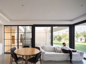 A modern living room with concrete and glass elements, featuring a white sofa and a round wooden dining table with black chai