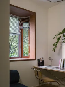 A modern home office featuring a wooden window with blinds, a desk with books and a chair, and green foliage outside the wind