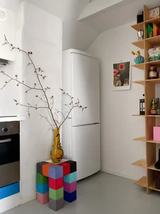 A minimalist industrial kitchen with wooden shelves, a white refrigerator, and colorful blocks under a vase on a colorful sta