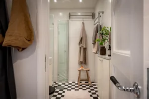 A minimalist bathroom featuring white tiled walls and floor, a glass shower stall, and hanging towels.