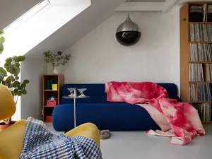 A modern minimalist living room features a blue sofa with a pink and red blanket, a yellow armchair with a blue and white bla