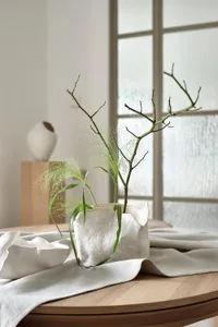 A modern minimalist white ceramic vase with green foliage and branches in a serene indoor setting.
