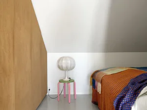 A minimalist bedroom featuring a colorful striped lamp on a pink and green stool, with an orange and blue patterned bedspread