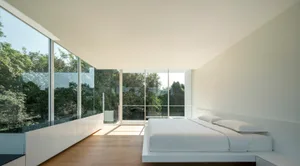 Minimalist bedroom with white furniture, large glass windows, and a view of green trees.