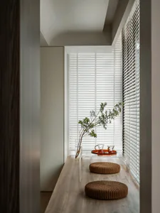 A minimalist living room with wooden furniture, clear glass vase, and white blinds.