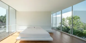 Minimalist bedroom with a single, white-floating platform bed with wooden flooring and large windows with a view of greenery 