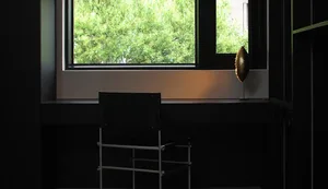 A minimalist interior design featuring a window with greenery outside, a black chair, and a metallic lamp on a dark shelf.
