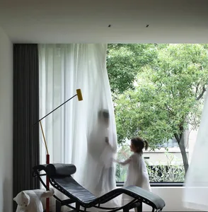 A contemporary living room featuring a sleek, black lounge chair with white curtains and a yellow desk lamp behind it, lookin