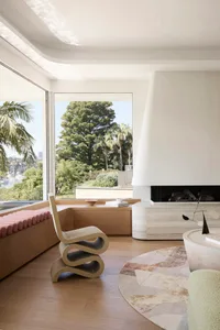 Modern living room with a wooden floor, white fireplace, and large windows overlooking greenery.