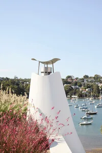 A modern metal sculpture with a curved top sits amidst flowers and plants near a harbor with sailboats.