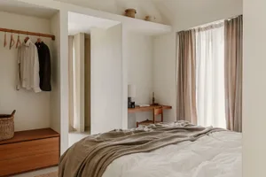 A minimalist bedroom with a bed covered in linen sheets and a wooden desk under a window with sheer curtains.