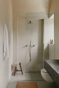 A minimalist concrete bathroom featuring a glass shower enclosure, stone sink, and wooden stool with a white towel.