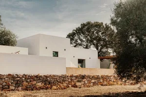 A minimalist white house with a concrete wall and a stone wall in the foreground, surrounded by trees.