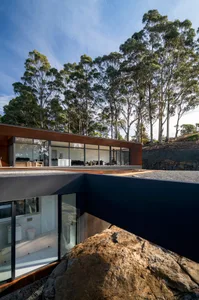 Modern residential building with large glass windows and dark metallic accents, surrounded by trees and rocks.
