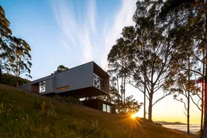 Modern residential house with a metal and glass exterior, situated on a grassy hillside with tall trees and a sunset.