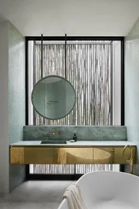 A modern bathroom with green marble counter and brass accents, featuring a round mirror and a large window.
