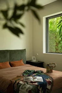 A modern bedroom featuring a bed with green headboard and colorful textiles, a small table with a vase and flower, and a wind