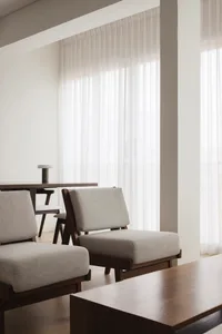 A modern minimalist living room featuring upholstered fabric chairs and wooden tables with sheer curtains allowing natural li