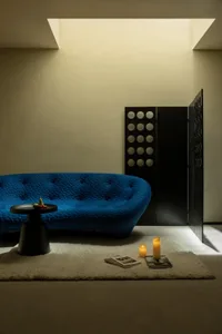 A minimalistic living room featuring a textured blue sofa, a small side table with a fruit bowl, and candles on a rug.